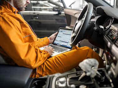 Mechanic in orange uniform using a laptop inside a car for diagnostic work.