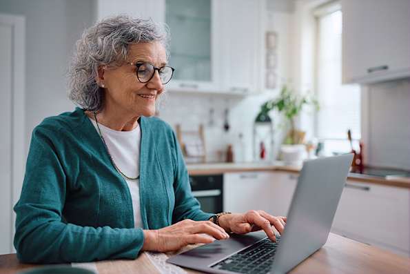 Elderly woman with glasses using a laptop at a kitchen table.