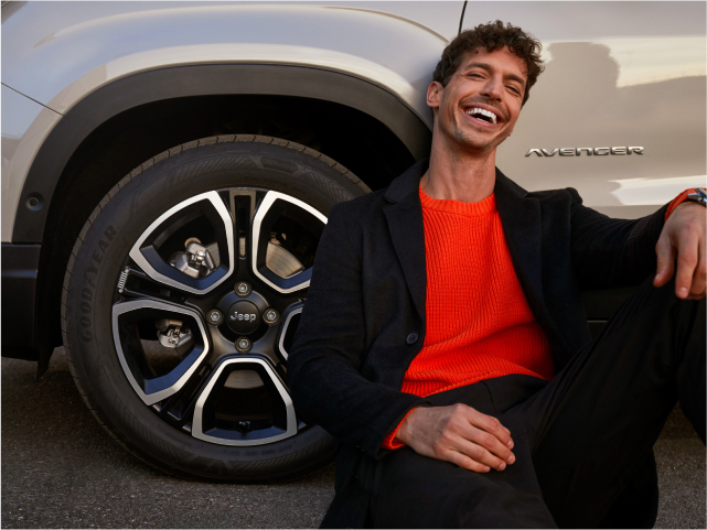 Man in orange jumper sitting beside a car wheel, smiling.