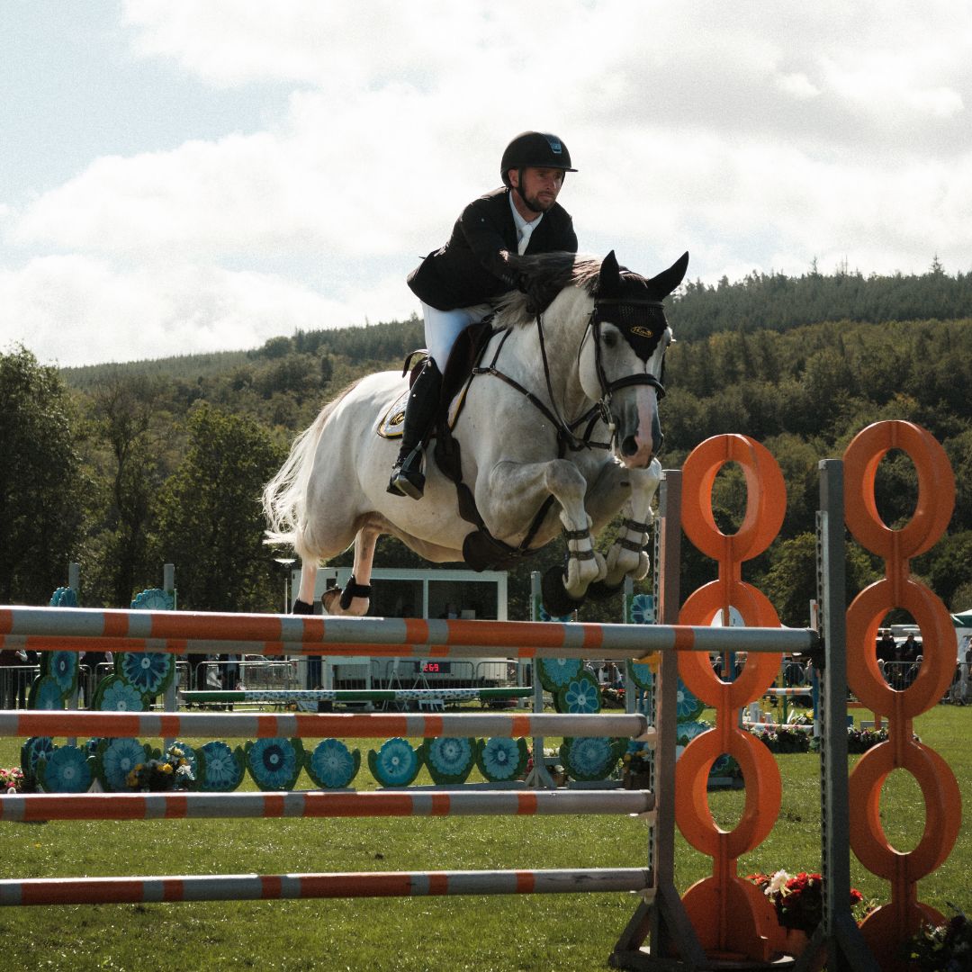 Rider and white horse jumping over an orange and white obstacle at an equestrian event.
